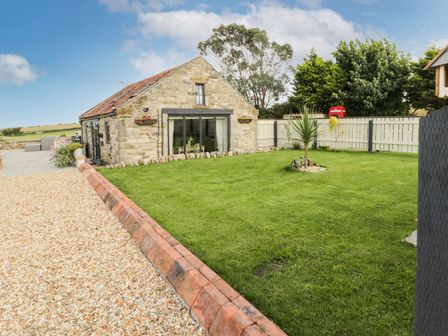 A stone house with a tiled roof next to a gravel pathway and a grass lawn with a small tree and a white fence at Cowshed Normanby near Flyingthorpe