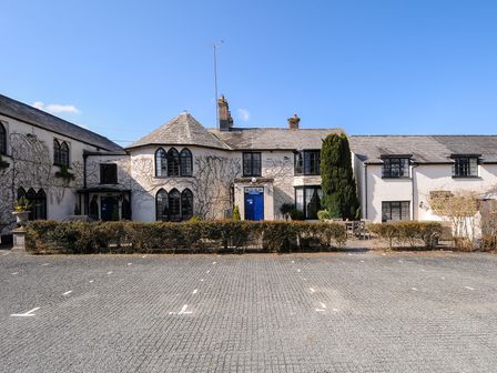 A stone paved parking area with marked spaces and a white building with multiple windows and blue doors behind a hedge at Alway in Lifton