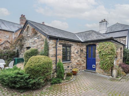 A stone cottage with a blue door and small window surrounded by bushes and a paved driveway at The Barn in Lifton