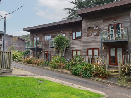 A wooden two story building with balconies and glass railings surrounded by plants and a road at Treburgett in Porthtowan