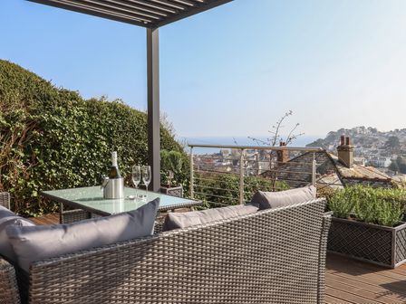 An outdoor patio with wicker furniture and a glass table with a bottle and two glasses overlooking a town and the sea at 15 Summerland Avenue in Dawlish