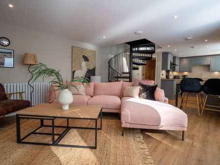 A living room with a pink sofa, coffee table with a plant, spiral staircase, and kitchen area with bar stools at Coral Cottage in Kessingland