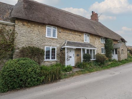 A stone thatched roof cottage with white framed windows and a porch along a quiet street at Cobblers Cottage in Langton Herring