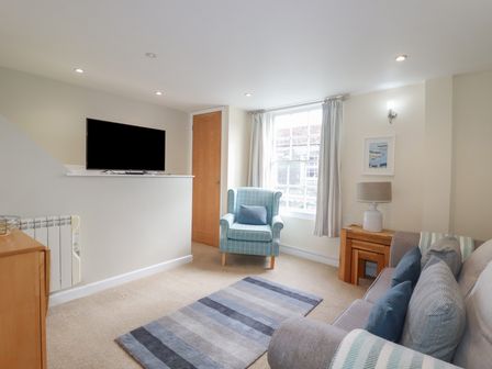 A living room with a blue armchair a sofa a wooden side table with a lamp and a television mounted on a half wall at Obby Oss in Padstow
