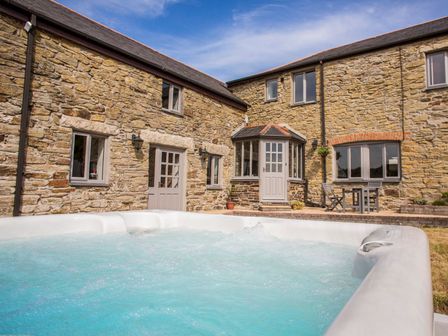 A stone house with multiple windows and doors with a bubbling hot tub and outdoor table and chairs at Five Elements Farmhouse in St Agnes