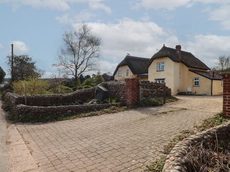 A driveway with a stone wall and a thatched roof house in the background at Badger's Den in Budleigh Salterton