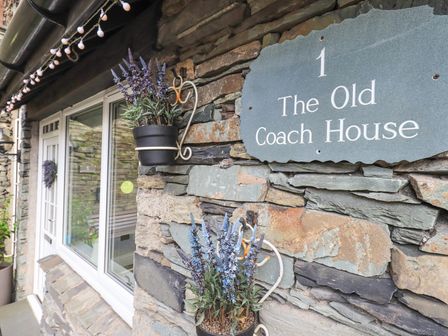 Exterior stone wall with a door and window and flower pots hanging near a sign that says 1 The Old Coach House Ambleside