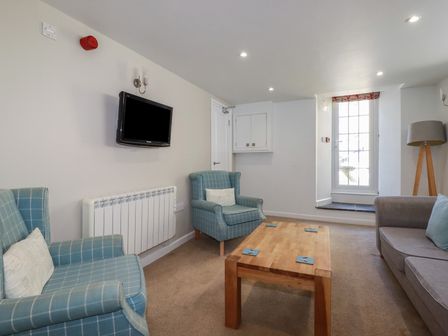 A small living room with two blue plaid armchairs a wooden coffee table a grey sofa a floor lamp a wall-mounted TV and a glass door at St Georges in Padstow
