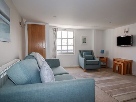 A living room with a blue sofa and armchair a wooden door a window wall-mounted TV and nesting wooden tables at Prideaux in Padstow
