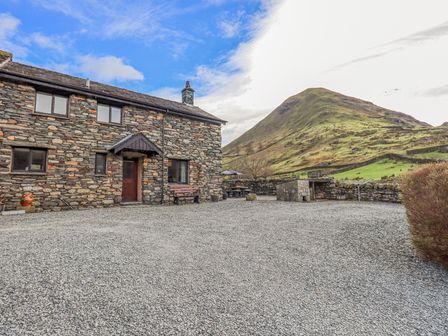 A stone house with a wooden door and bench on a gravel yard with a mountain in the background at Low Wood View in Hartsop
