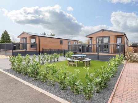 Two brown mobile homes with fenced decks and a picnic table on artificial grass surrounded by plants at Blossom Vale Lodge in Evesham