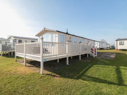 A mobile home with a wooden deck and stairs on a grassy lot at Rose Bank D8 in Primrose Valley