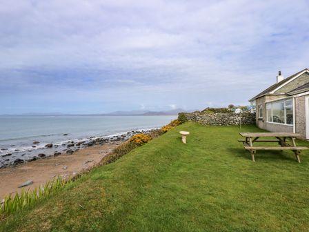 A grassy backyard with a picnic table and stone birdbath beside a house overlooking a rocky beach and ocean at Tegawelon in Llandanwg near Harlech