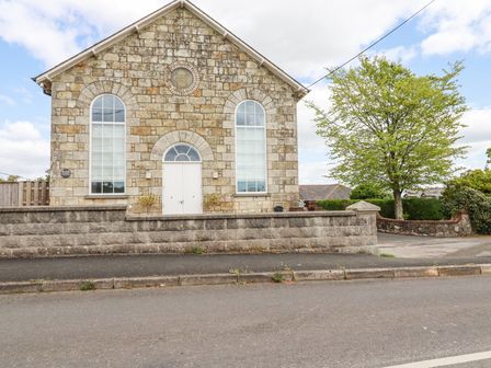 The exterior of a stone building with arched windows and a white door behind a stone wall and a tree at Trelowth Chapel in Polgooth