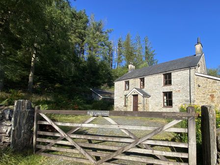 A stone cottage behind a wooden gate with surrounding trees at Cae'n Y Coed Cottage in Penrhyndeudraeth