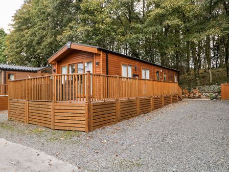A wooden cabin with a fenced deck and gravel driveway surrounded by trees at Water's Reach White Cross Bay Holiday Park near Troutbeck Bridge