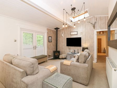A living room with two beige sofas a wall-mounted television a small stove and double doors at Serenity Skiptory Howe 10 White Cross Bay near Windermere
