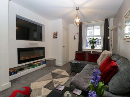A living room with a large grey sectional sofa with red cushions a wall-mounted television above a fireplace and a window with closed curtains at Colliers Cottage in Cinderford