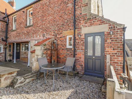 An outdoor patio area with a round glass table and two chairs next to a brick building with a dark door at Seafield Granary in Alnmouth