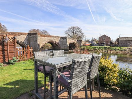 An outdoor seating area with a glass table and wicker chairs by a river near a stone bridge at Beckside Cottage in Penrith