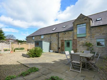 A stone house with green door and windows a patio with wooden table and chairs and a garden shed at Croft Cottage in Alnwick