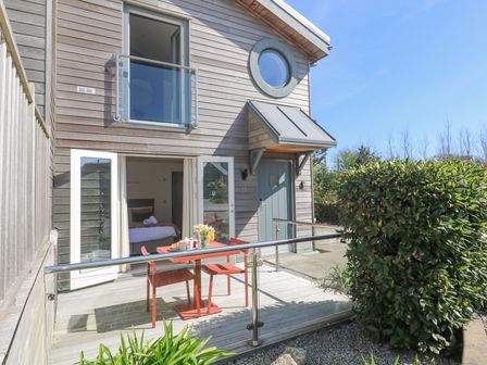 An outdoor patio with a red table and chairs outside a wooden house with open doors showing a bedroom at Azure Retreat at Una Carbis Bay near St Ives