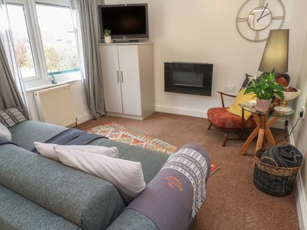 A living room with a blue sofa pillows a patterned rug a white cabinet with a TV wall clock a red chair a glass side table with a plant and a lamp at Ty Metheven in Camborne