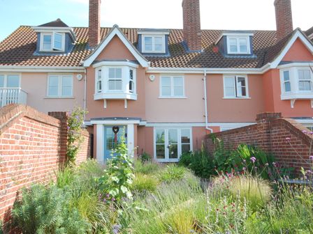 A garden with plants and flowers in front of a terraced house with pink walls and a blue door at Sea Crest in Aldeburgh