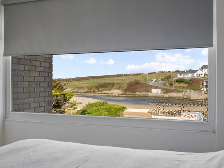 A window with a gray roller blind partially down showing a river, grassy hill, road, and buildings outside at 5 Mermaid Court in Newquay