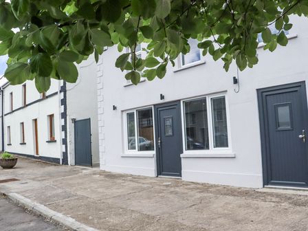 The exterior of a white building with dark gray doors and windows and a tree with green leaves above on a concrete pavement at Daly's Place in Ballyporeen County Tipperary