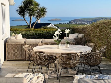 An outdoor patio with wicker chairs around a round wooden table and a wicker sofa set with beige cushions overlooking the sea at Trem Ynys 3 bed in Abersoch