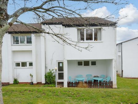 A white two-story building with a covered patio and blue outdoor furniture on grass at Honipine House Callington near Gunnislake