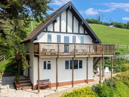 A two story house with a wooden balcony and outdoor seating on a hill with trees and a grassy field at Seascape Cottage in Beer