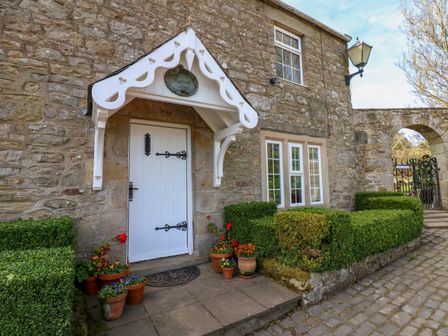 An entrance door with potted plants under a white porch at the front of a stone cottage with hedge and a cobbled pathway at Pagecroft Cottage in Haydon Bridge