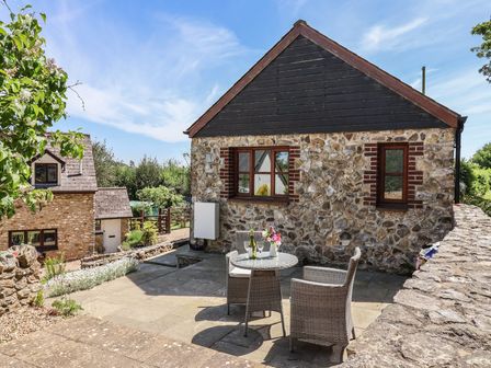A stone patio with a round table and two wicker chairs outside a stone and wood building at Megs Roost Shute near Colyton