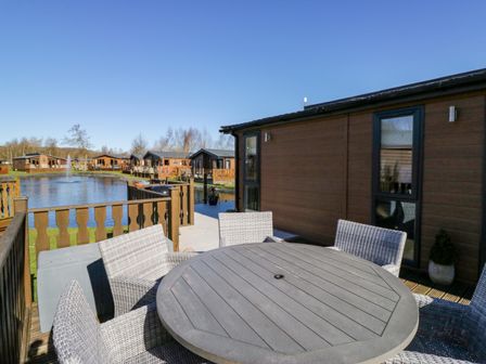 A patio with round table and chairs next to a wooden lodge and a pond at Coniston View Lodge in Warton near Carnforth