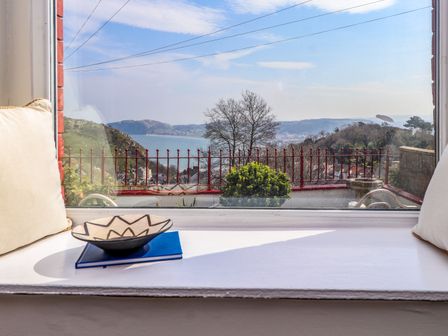 A window seat with cushions and a bowl on a book overlooking a view of a railing trees hills and a body of water at 4 Bay View Terrace in Llandudno