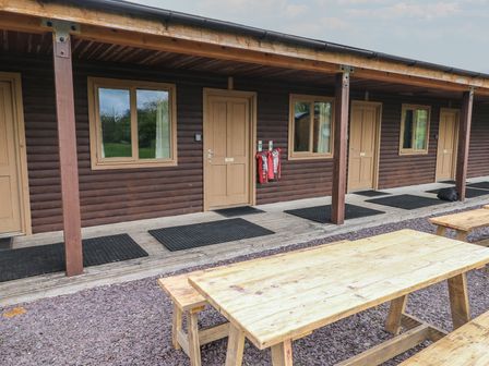 An outdoor seating area with wooden picnic tables in front of a row of wooden cabins with doors and windows at Alder in Cowbridge