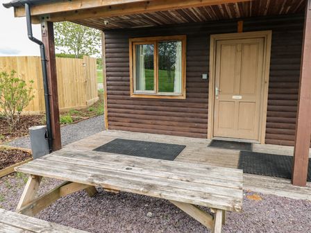 An outdoor wooden picnic table in front of a cabin porch with a window and a door labeled Oak at Oak in Cowbridge