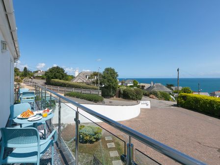 A balcony with blue chairs and tables overlooking a street with houses and the sea in the background at Skysail in Carbis Bay