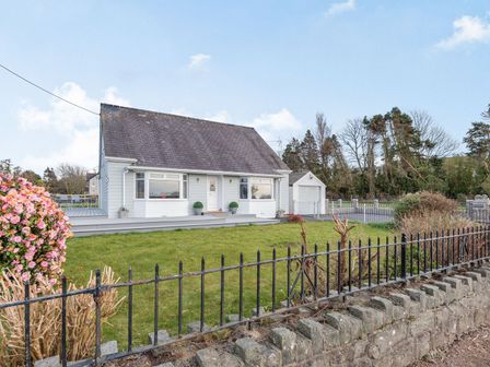 A house with a sloped roof and a front yard with grass and flowers behind a metal fence at Bryn Derw in Caernarfon
