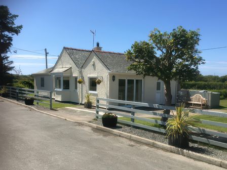 A white single-story house with a gray fence plants and a tree outside at Plas Lodge in Llanfaelog