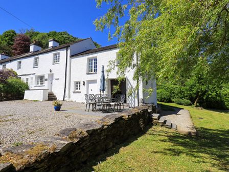 An outdoor patio with table and chairs outside white cottages surrounded by trees and grass at Great Hartbarrow Farm Cottage in Cartmel Fell near Windermere