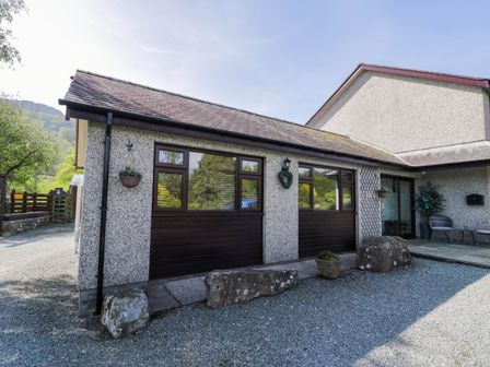 The exterior of a single story building with two large dark wooden windows rocks gravel in front and outdoor seating at Gilfach Bach in Dolgellau