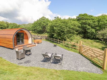 An outdoor area with a wooden pod cabin two chairs and a small table a hot tub and a fenced grassy area at Eirlys in Bontddu near Dolgellau