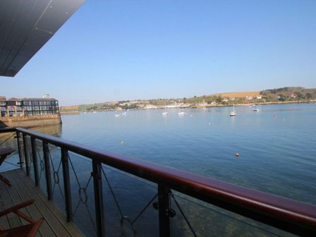 A balcony with wooden chairs overlooking a body of water with boats and a distant shoreline at Tobys Quay in Falmouth