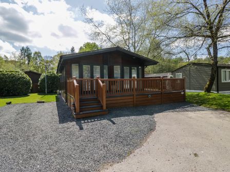 A wooden lodge with a deck and stairs surrounded by gravel and grass with trees nearby at Robins Lodge in White Cross Bay near Troutbeck Bridge