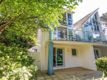 An exterior view of a house with a glass balcony and patio surrounded by trees and bushes at 6 Red Sails in Sandbanks