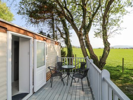 A deck with a round glass table and four black chairs outside a mobile home near a grassy field at No 29 Wyndham Hall in Cockermouth