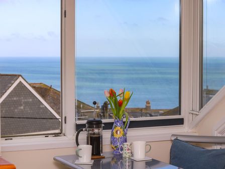 A table with a coffee press, three mugs, and a vase of tulips by a window overlooking the sea and rooftops at Breakers Point in St Ives